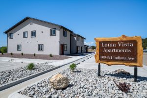 Colorado Mountain Property and Homes for Sale - A modern, two-story apartment building with a wooden sign in front reading "Loma Vista Apartments, 809-821 Indiana Ave." offers units for rent. The area is landscaped with rocks and small plants under a clear blue sky.