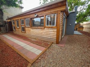 Colorado Mountain Property and Homes for Sale - A wooden shed with multiple windows sits beside a gravel and paved walkway, surrounded by red mulch and trees in the fenced backyard of 118 E 7th St, Walsenburg CO, available for rent.