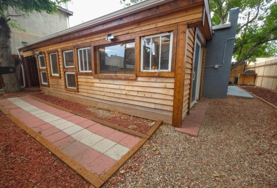 Colorado Mountain Property and Homes for Sale - A wooden shed with multiple windows sits beside a gravel and paved walkway, surrounded by red mulch and trees in the fenced backyard of 118 E 7th St, Walsenburg CO, available for rent.